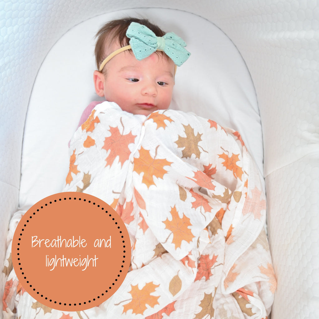 A baby girl snuggled under the 100% Muslin Cotton Fall Leaves Swaddle Blanket - 47" x 47", experiencing warmth and comfort in the beautiful embrace of autumn.