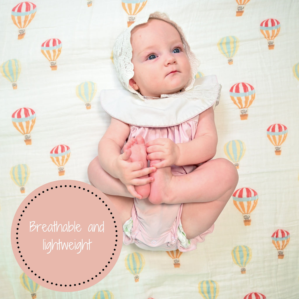 A delighted baby girl with a radiant smile, set against the backdrop of the Up, Up, and Away 100% Muslin Cotton Swaddle Blanket - 47" x 47", spreading joy and the playful essence of hot air balloons.