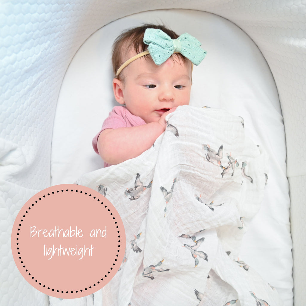A happy baby girl with a radiant smile, comfortably cocooned in the Mallard Magic Muslin Swaddle Blanket - 47" x 47", resting peacefully in her bassinet, surrounded by the charm of mallard ducks.