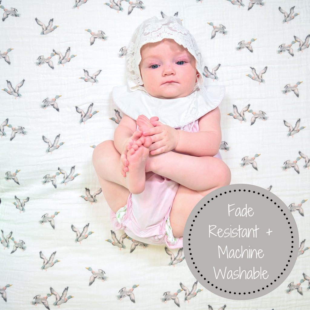 A delighted baby girl with a radiant smile, set against the backdrop of the Mallard Magic Muslin Swaddle Blanket - 47" x 47", spreading joy and the playful essence of mallard ducks.