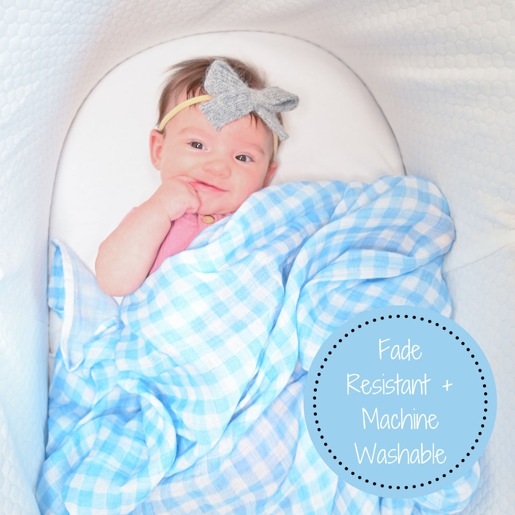 A joyful baby girl with a radiant smile, comfortably cocooned in the Classic Comfort Blue Gingham Muslin Swaddle Blanket - 47" x 47", resting peacefully in her bassinet, embraced by timeless comfort.