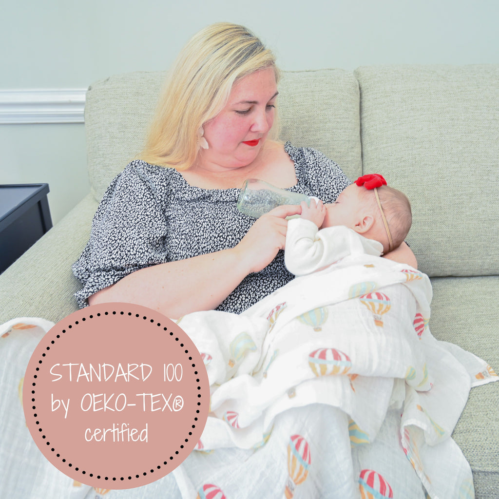 A caring mom holding her baby, wrapped in the Up, Up, and Away Muslin Swaddle Blanket - 47" x 47", while nurturing the baby with a loving bottle of milk.