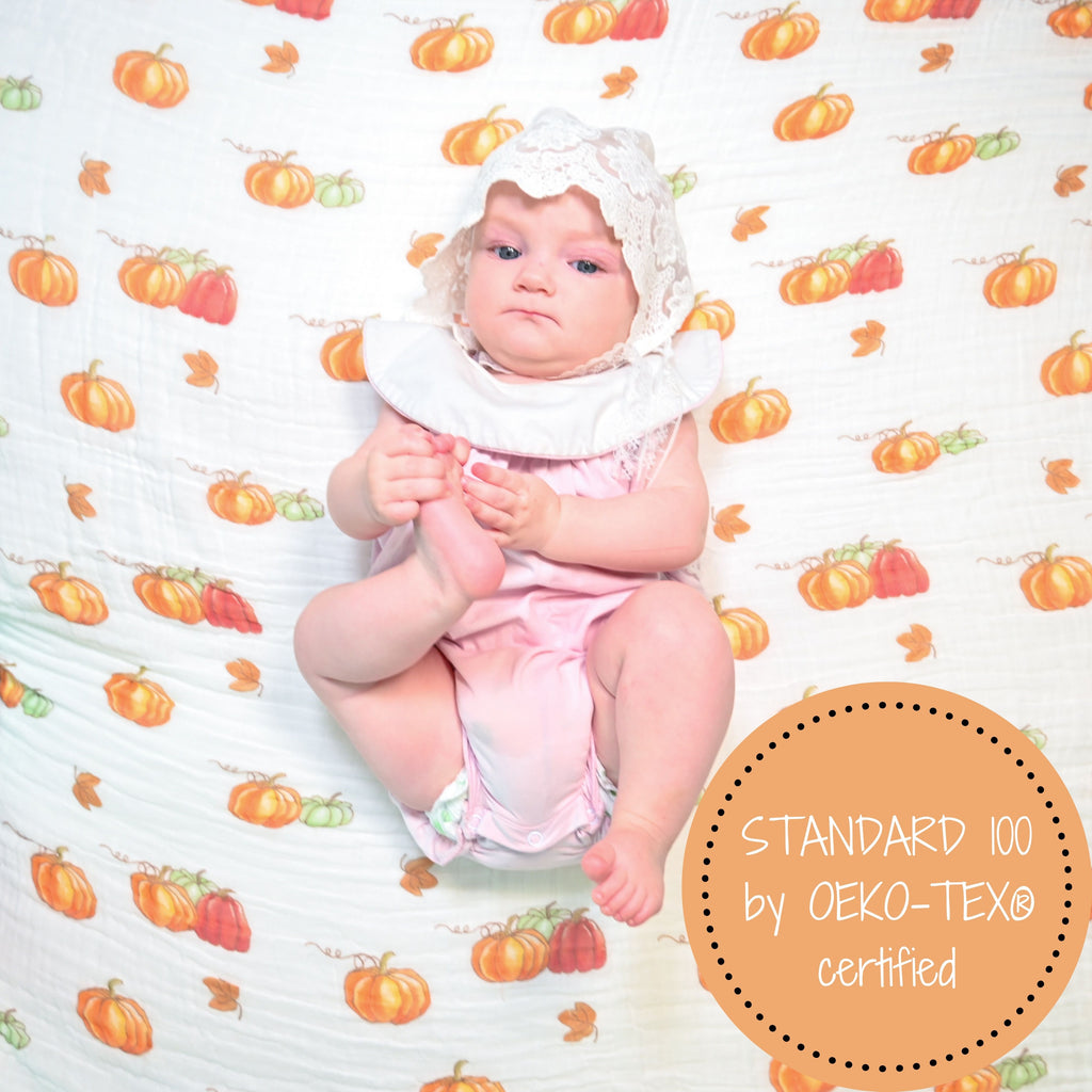 A happy baby girl with a radiant smile, framed by the 100% Muslin Cotton Pumpkin Swaddle Blanket - 47" x 47", exuding joy and warmth with the pumpkin-themed backdrop